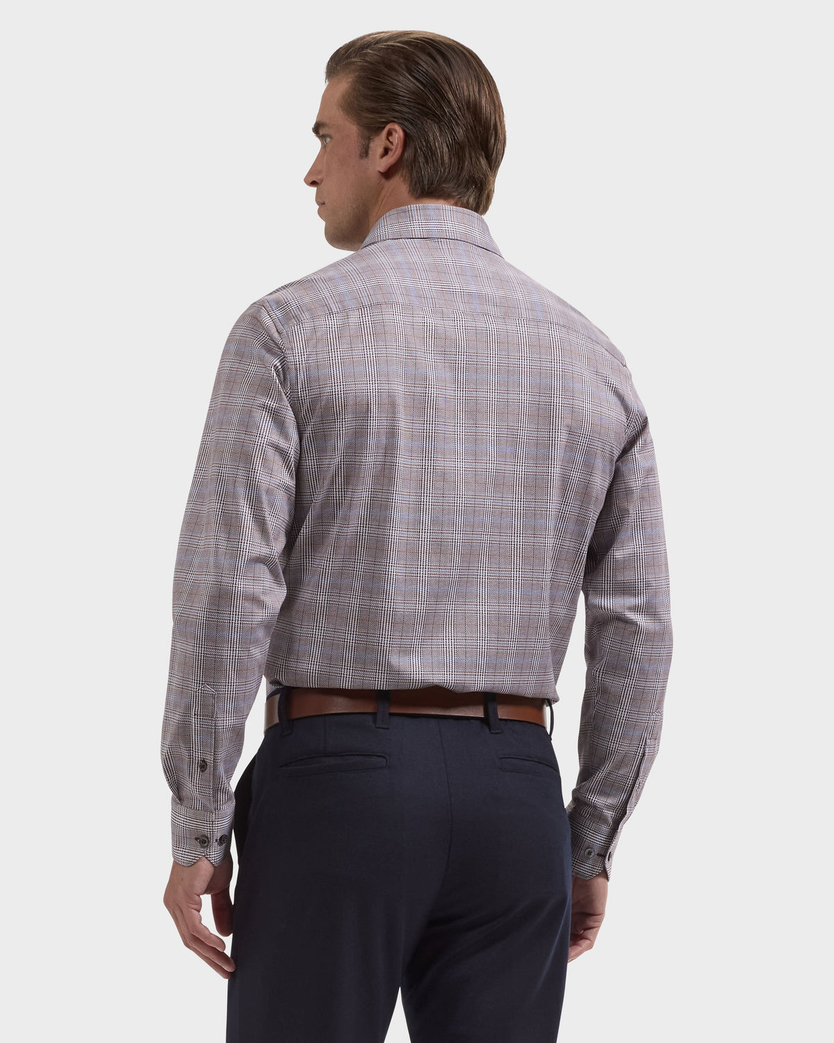 A man shown from the back wearing a long-sleeve plaid button-down shirt in muted gray, beige, and blue tones, paired with dark navy trousers and a brown belt, against a light gray studio background.