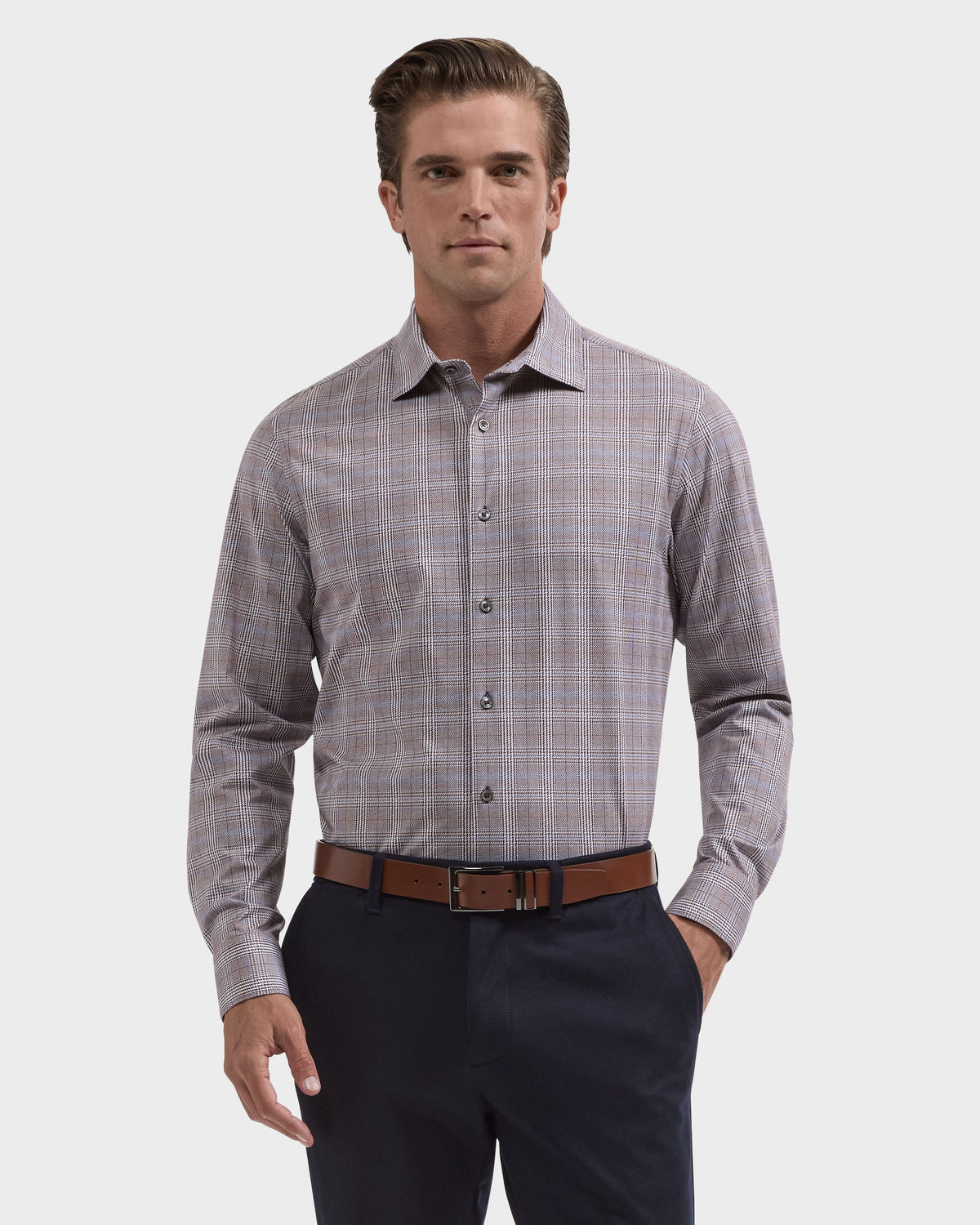 A man modeling a long-sleeve button-down dress shirt with a fine plaid pattern in muted gray and pink, paired with dark trousers and a brown belt, against a light gray studio background.