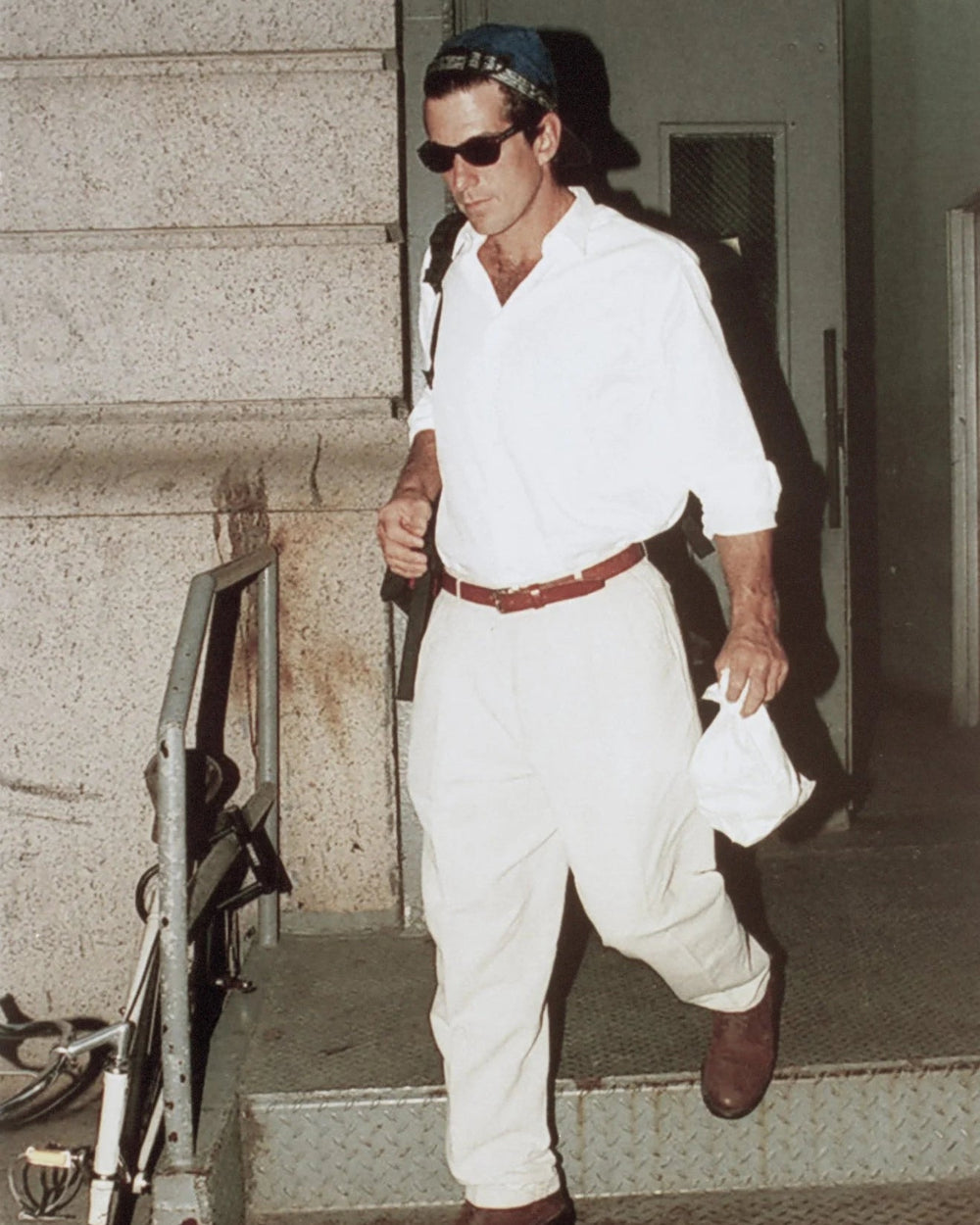 A man wearing a white long-sleeve shirt, white pants, a brown belt, and brown shoes walks down concrete stairs while wearing dark sunglasses and a dark cap.