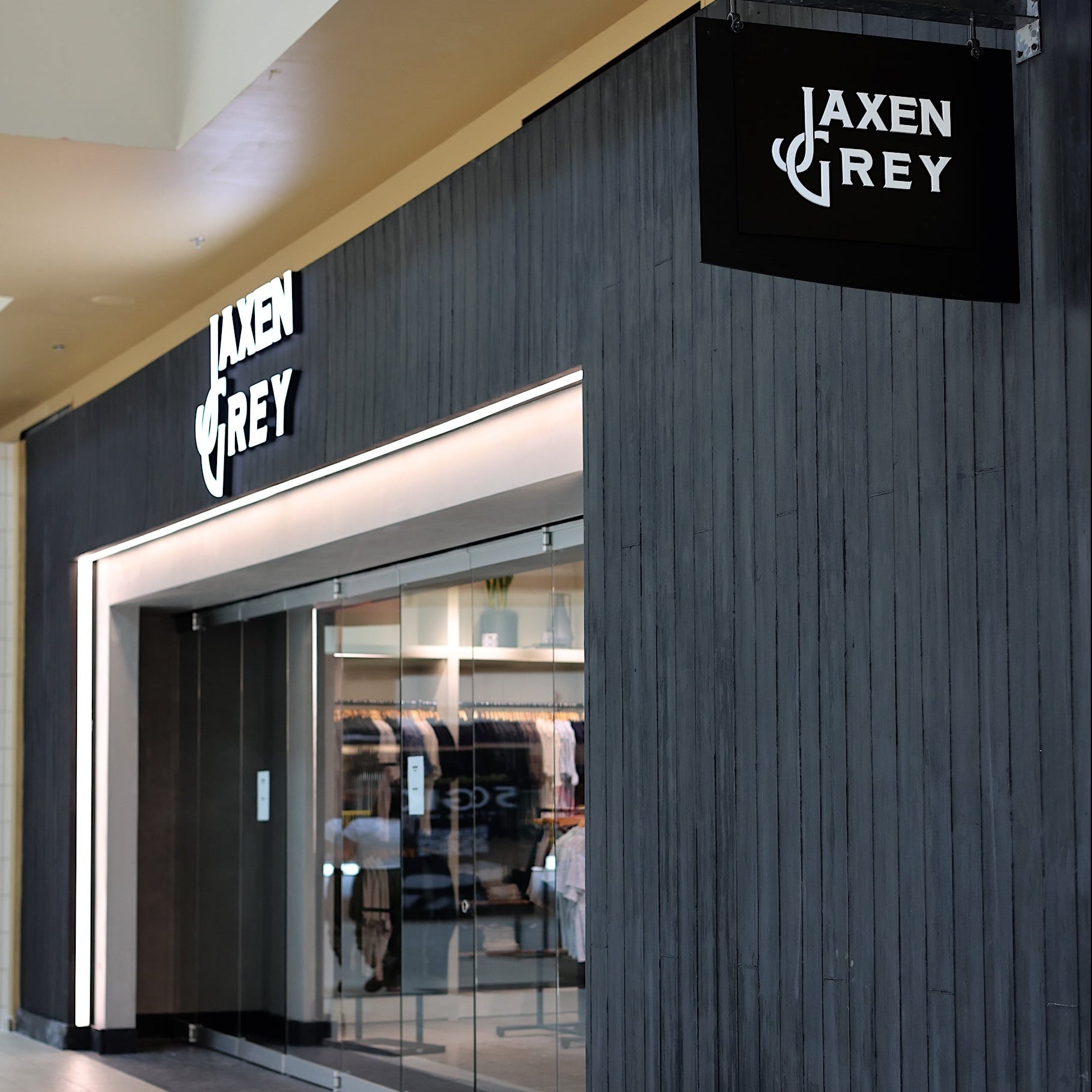 Jaxen Grey clothing store storefront with dark wood paneling, glass entrance, and a black hanging sign on a mall corridor.