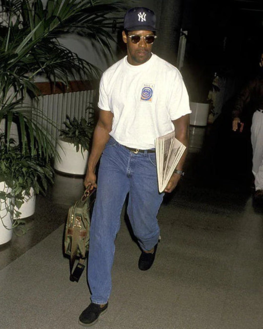 A man walking through an airport terminal wearing a white t-shirt, blue jeans, a New York Yankees baseball cap, sunglasses, and carrying a green bag and a newspaper.