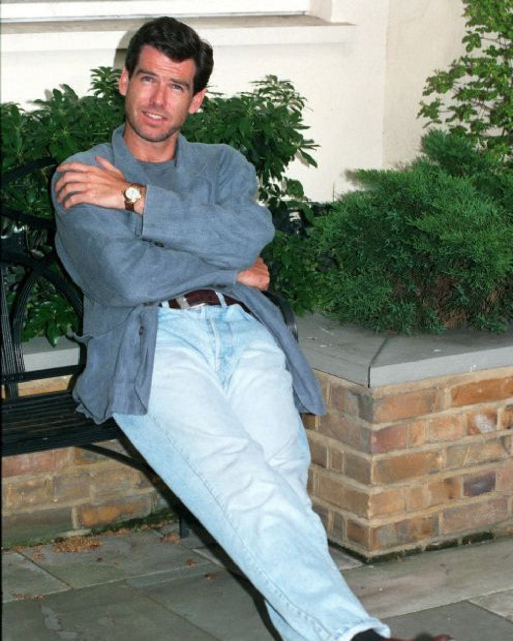 A man sitting on a black metal bench outdoors, wearing a blue-gray long-sleeve button-up shirt and light blue jeans, with green plants and a brick planter in the background.